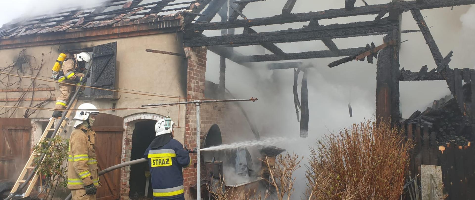 Dwóch strażaków podających prąd wody do wnętrza zniszczonego przez ogień, zadymionego budynku. Z lewej strony strażak wchodzący do budynku po drabinie przez otwór na piętrze.