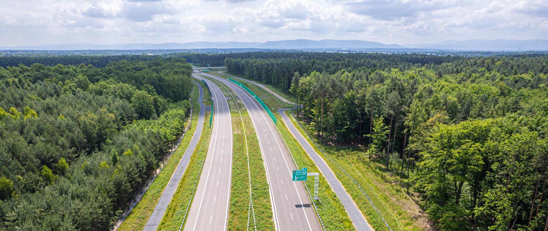 Szeroka droga ekspresowa S1 niedługo przed zakończeniem prac, w tle widok na rozciągające się pasmo Beskidu Żywieckiego. To znak, że zbliżamy się do południowych krańców województwa śląskiego, a tym samym Polski. Wokół drogi zachowany został drzewostan, co pozwala na odniesienie wrażenia, iż podróżujemy niemal w lesie.