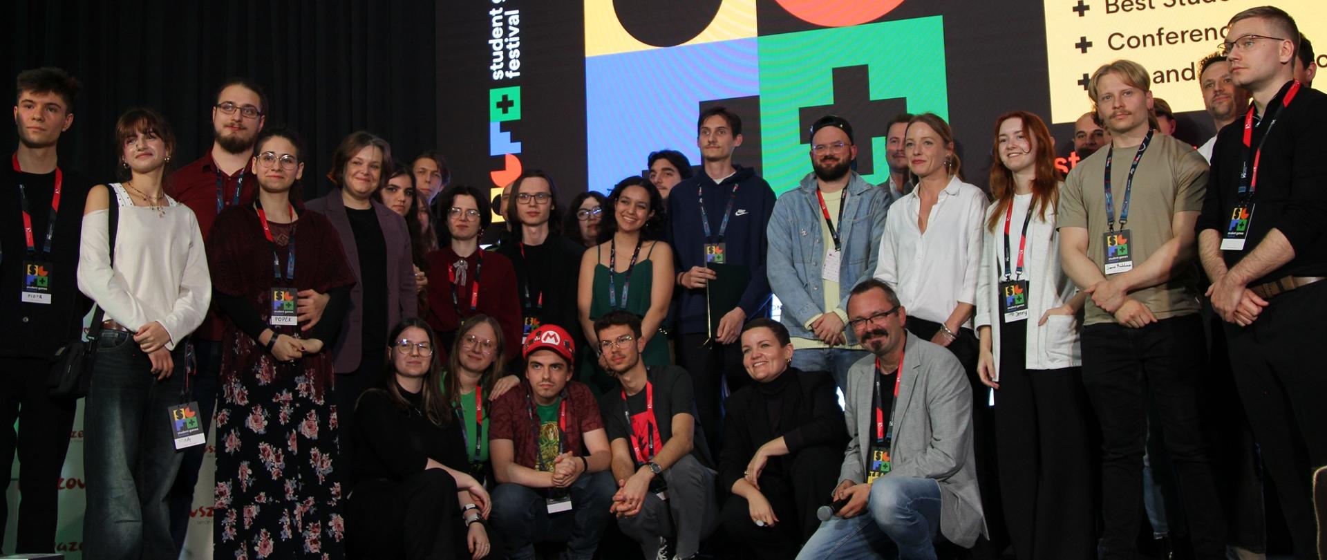 Na sali pod ścianką z napisem Student Games Festival stoi i klęczy w kilku rzędach duża grupa osób, z pośrodku grupy wiceminister Zioło-Pużuk.