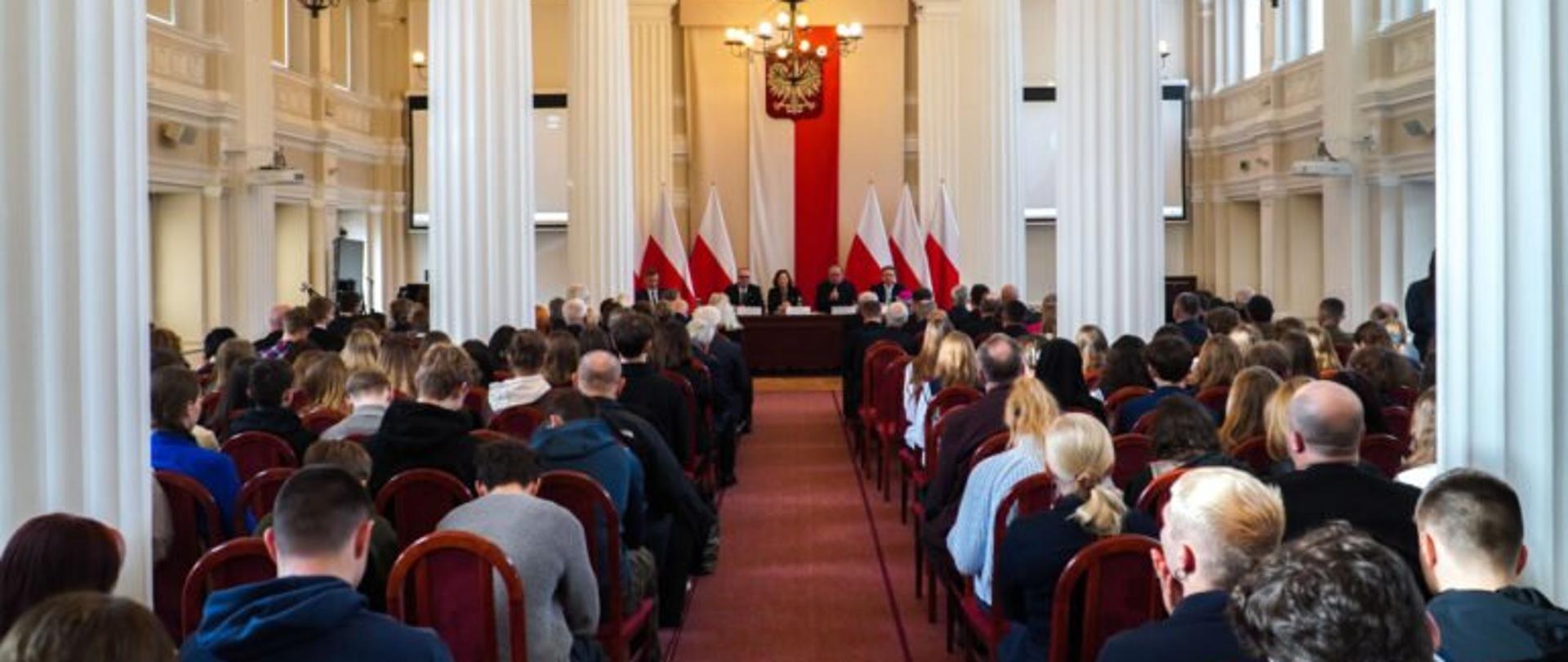 Uczestnicy konferencji dotyczącej arcybiskupa Tokarczuka zgromadzeni w sali kolumnowej Podkarpackiego Urzędu Wojewódzkiego w Rzeszowie, zajmujący miejsca siedzące przodem do stołu prezydialnego, za którym siedzą prelegenci