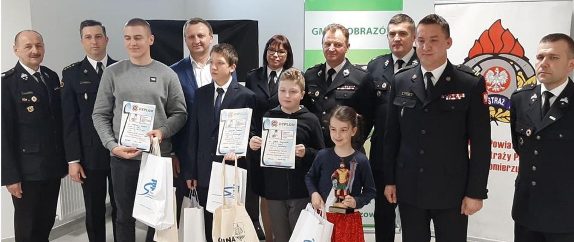 Zdjęcie wykonane w pomieszczeniu. Na zdjęciu widać stojących w pierwszym rzędzie zwycięzców OTWP szczebla powiatowego którzy trzymają dyplomy. W drugim rzędzie stoją strażacy ubrani w mundury wyjściowe oraz starosta sandomierski.