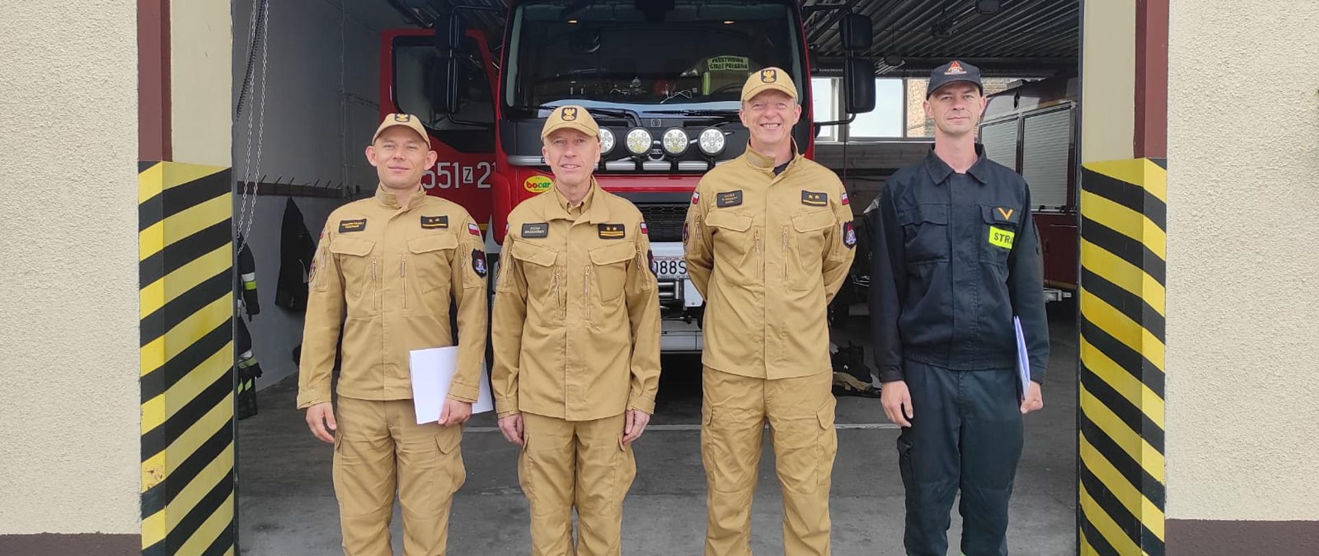 Zdjęcie przedstawia czterech strażaków na tle samochodów ratowniczo-gaśniczego stojącego w garażu.