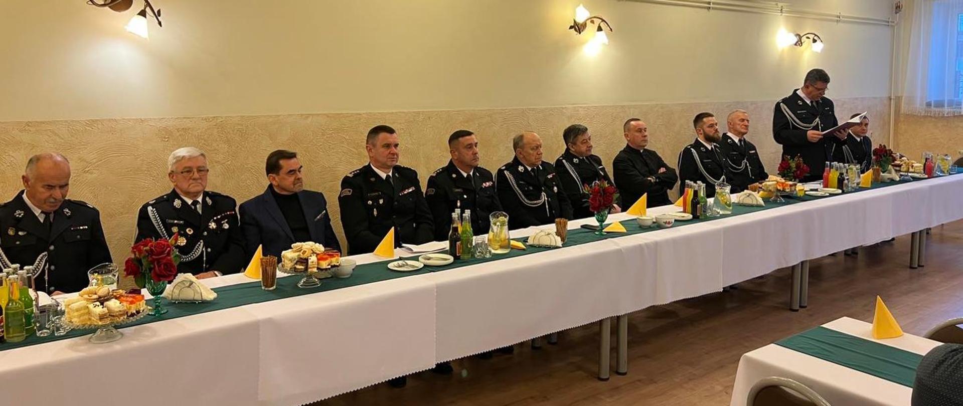 Zdjęcie zrobione w pomieszczeniu. Przy stole siedzi dwunastu mężczyzn, uczestników zebrania. Część z nich ubrana jest w mundury wyjściowe Państwowej Straży Pożarnej, a część w mundury Ochotniczej Straży Pożarnej. Jeden z mężczyzn stoi i czyta dokument.