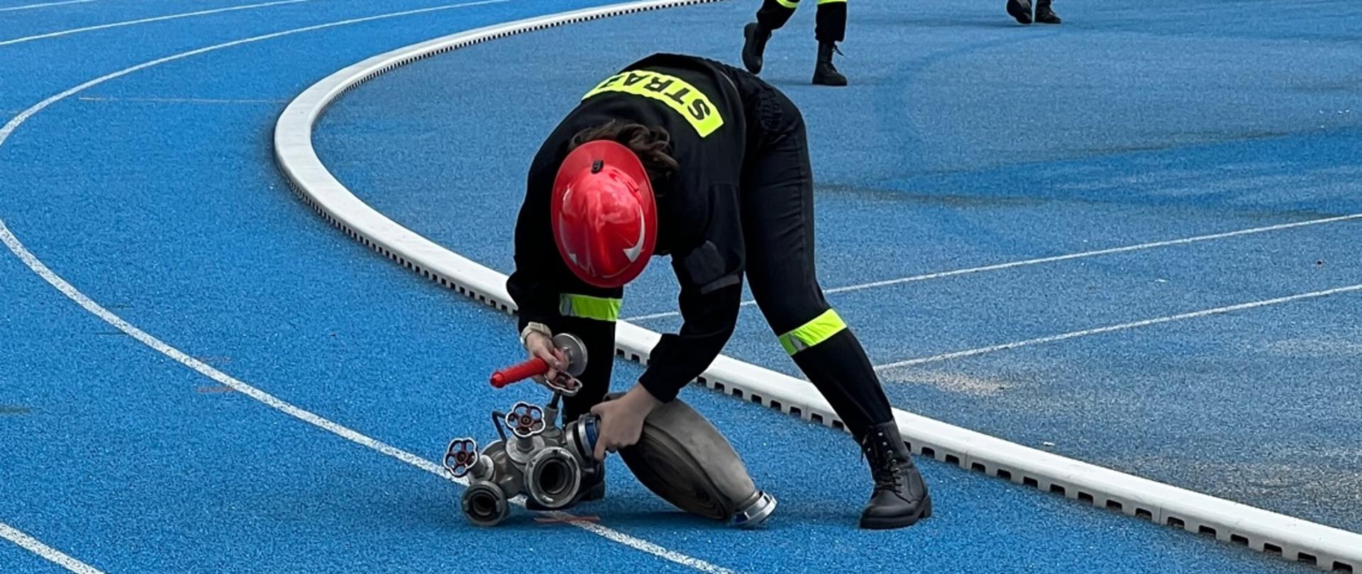Strażak OSP w czarnym ubraniu koszarowym oraz czerwonym hełmie podpina wąż do rozdzielacza, który stoi na niebieskim tartanie. W tle trzech innych strażaków.