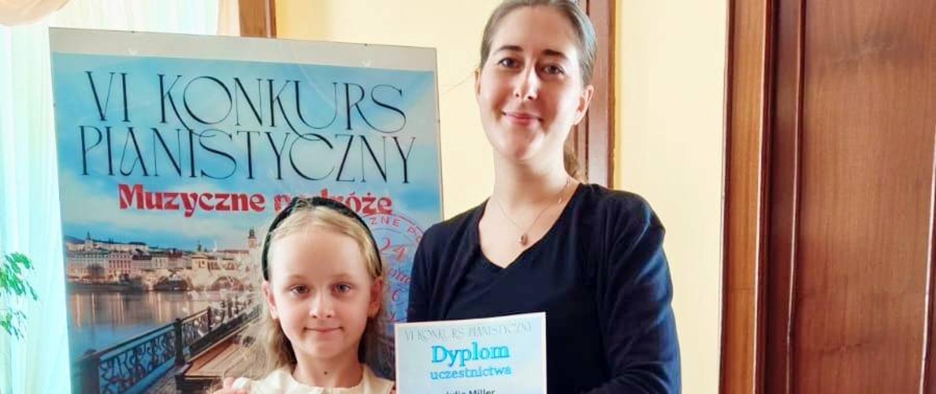 Pani Katarzyna Dzierzgowska pozuje wraz z uczennicą. Dziewczynka trzyma dyplom. W tle sztaluga z logo konkursu.