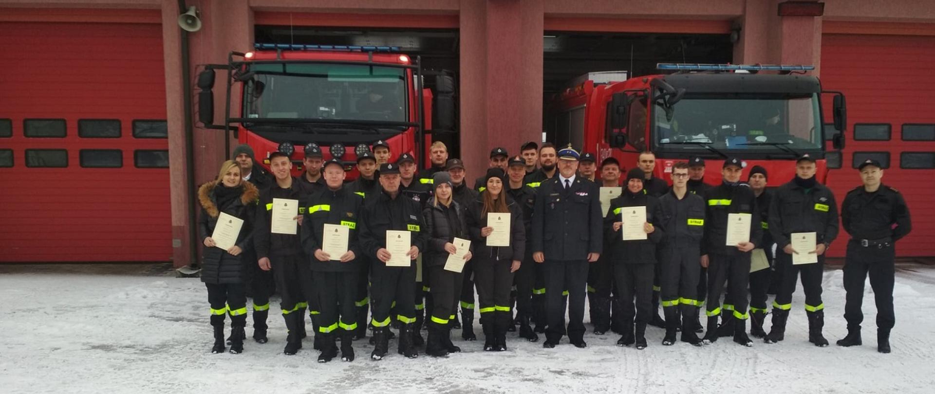 Zdjęcie obrazuje strażaków PSP i OSP ze świadectwami w dłoniach w tle pojazdy pożarnicze i budynek.