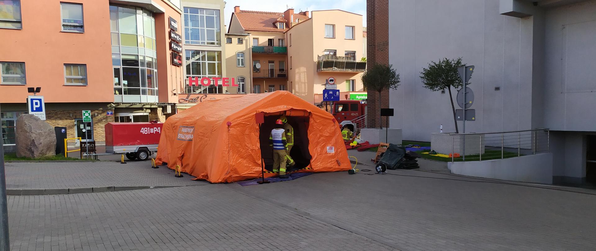 Fotografia przedstawia namiot strażacki w kolorze pomarańczowym stojący przed galerią handlową podczas ćwiczeń.