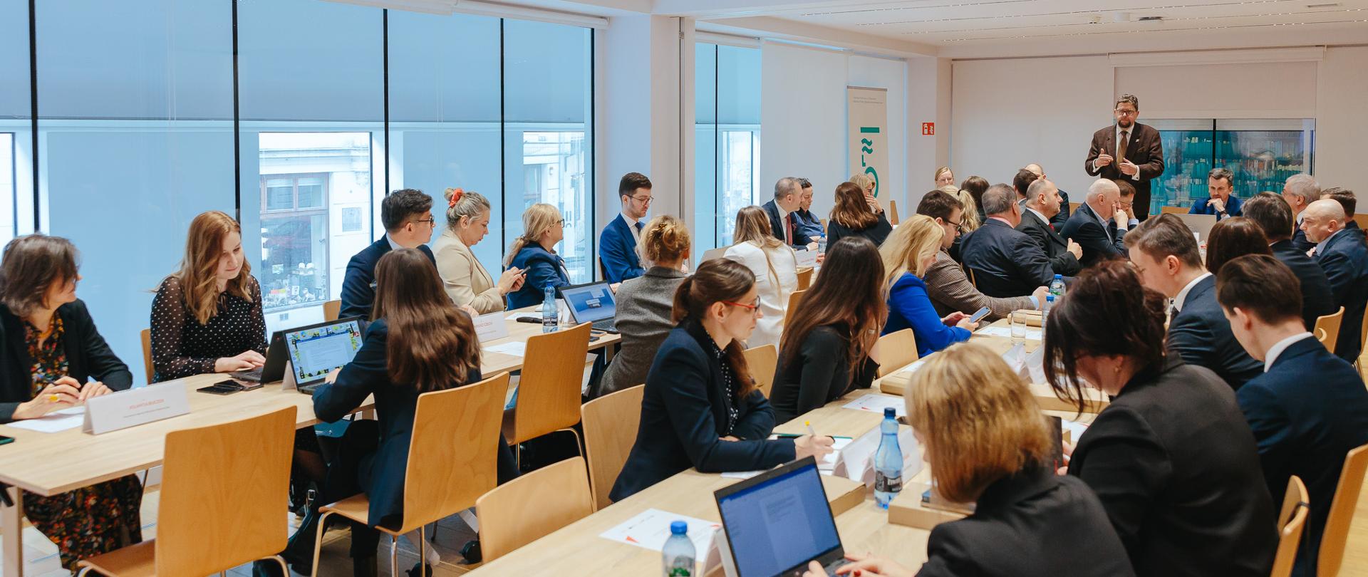 Widok z tyłu pod kątem na salę, przy ustawionych w czworokąt stołach siedzą ludzie, w głębi stoi wiceminister Szeptycki i mówi.