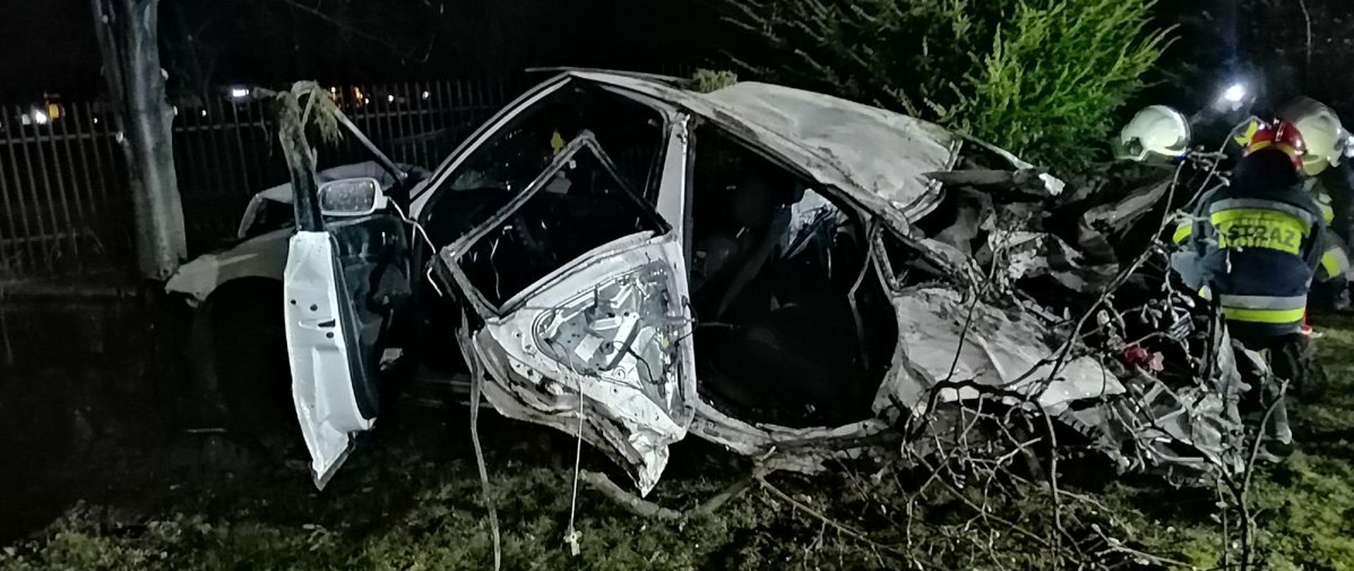 Zniszczony samochód koloru białego na trawie. Obok samochody prowadzą dzialania.