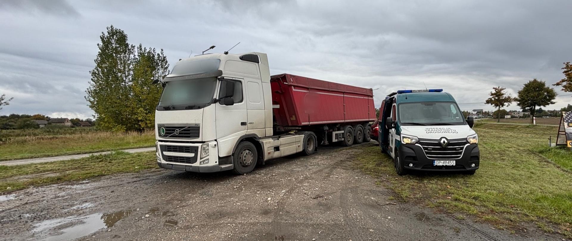 Biała ciężarówka z naczepą w czerwonym kolorze stoi na poboczu polnej drogi obok pojazdu służbowego Inspekcji Transportu Drogowego. W tle widać drzewa i pochmurne niebo.