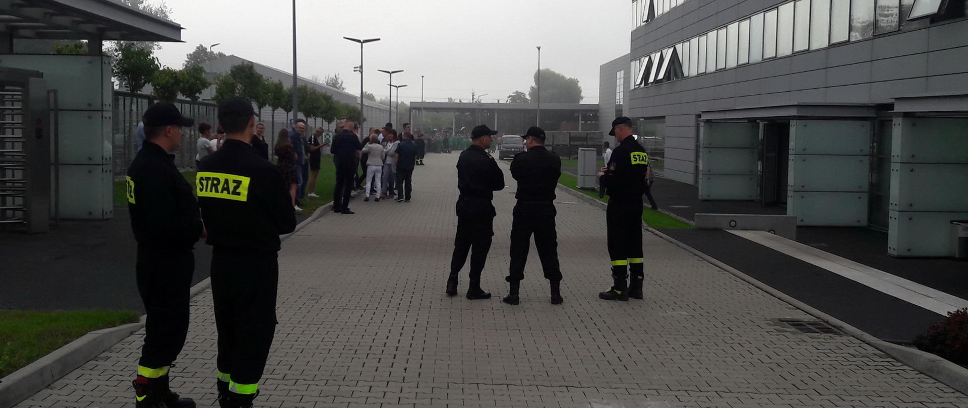 Na pierwszym planie widoczna jest grupa strażaków, za nimi widoczni są ewakuowani ludzie, jeden ze strażaków zapisuje coś w notesie, pozostali rozmawiają ze sobą.