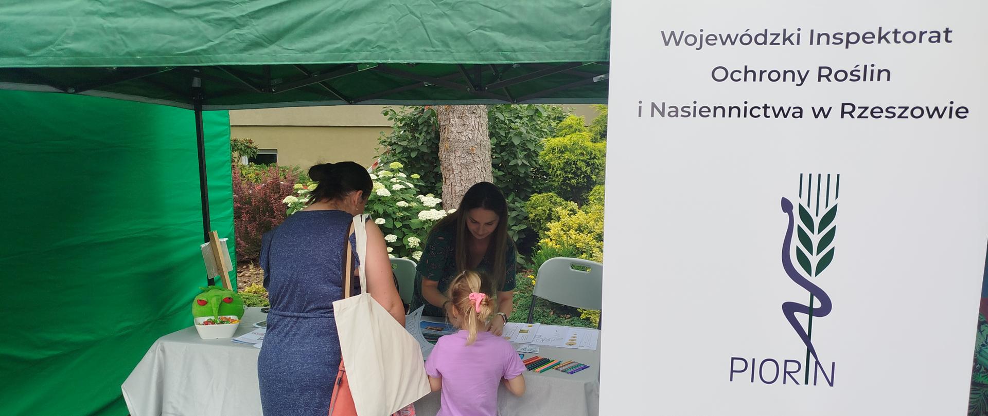 Kobieta i dziecko stoją pod zielonym namiotem pod którym jest stolik i porozkładane na nim gadżety i kredki. Na stołem stoi kobieta która pokazuje coś dziewczynce. Po prawej stronie stoi biało-granatowy baner logiem instytucji. 