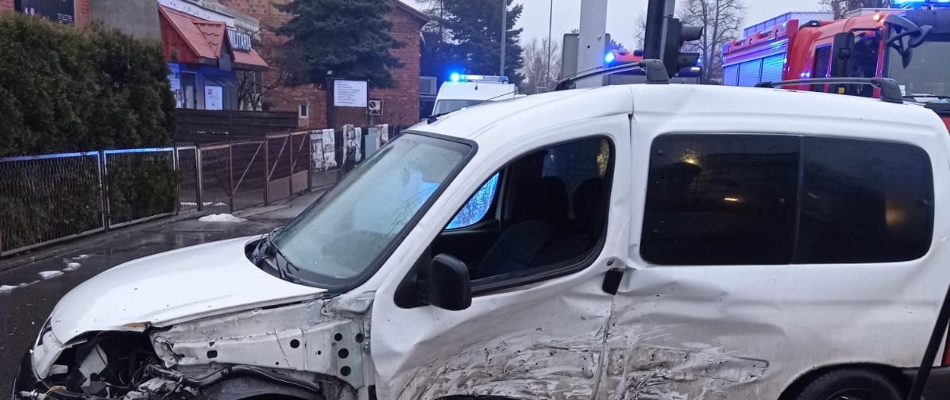 Na zdjęciu widać uszkodzony pojazd osobowy. Uszkodzony bok od kierowcy. Pojazd w kolorze białym. Z tyłu budynki mieszkalne położone przy ulicy Stawiszyńskiej. Prawy górny róg pojazdy gaśnicze z włączoną sygnalizacją ostrzegawczą. Pojazdy kolor czerwony. Niebo zachmurzone. Pora zimowa.