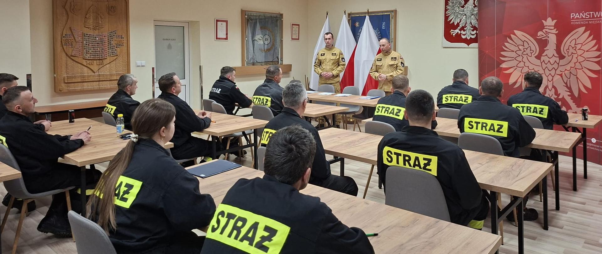 strażacy ubrani czarne ubrania koszarowe przed egzaminem teoretycznym w Sali szkoleniowej, w tle funkcjonariusze z komisje egzaminacyjnej ubrani w piaskowe mundury, ponadto godło i flaga Polski