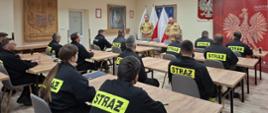strażacy ubrani czarne ubrania koszarowe przed egzaminem teoretycznym w Sali szkoleniowej, w tle funkcjonariusze z komisje egzaminacyjnej ubrani w piaskowe mundury, ponadto godło i flaga Polski