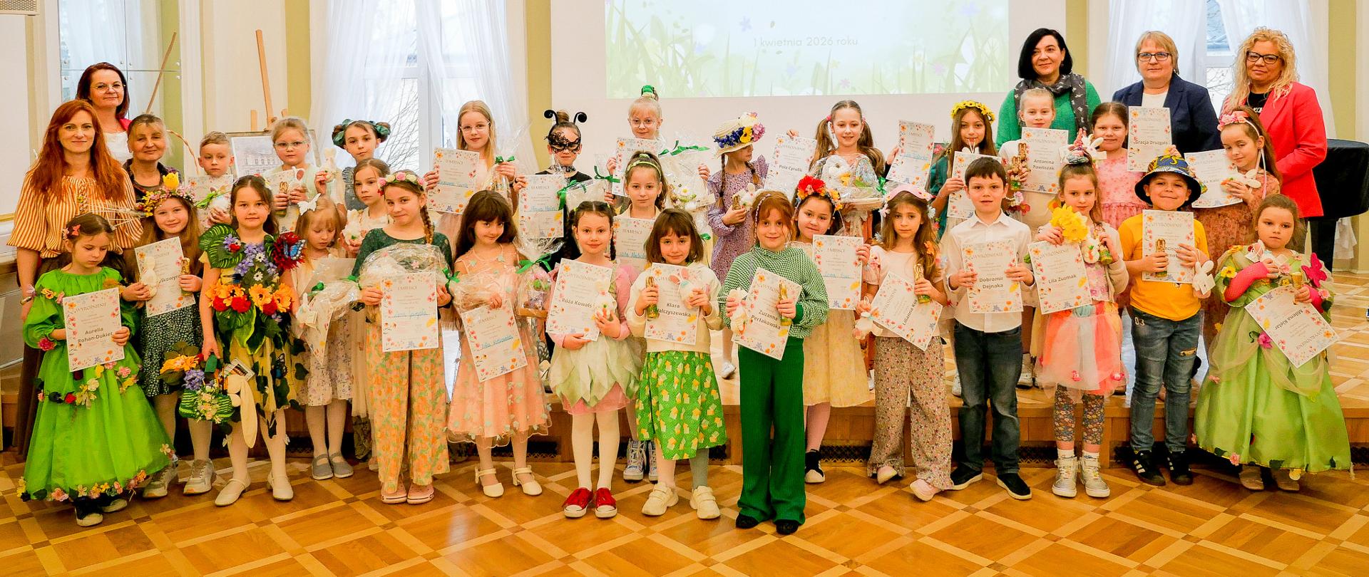 Grupa kilkudziesięciu dzieci stoi na scenie w jasnej, eleganckiej sali, trzymając dyplomy i drobne nagrody. Wiele z nich ma kolorowe, wiosenne stroje oraz ozdoby z kwiatów. Za nimi stoją nauczycielki lub opiekunki. Na ścianie w tle wyświetlony jest napis: „Szkolny Konkurs Recytatorski ‘Wiosna, ach to ty’” wraz z datą. Wnętrze zdobią wysokie okna, jasne ściany i elementy architektoniczne, a nad sceną widoczny jest polski orzeł.