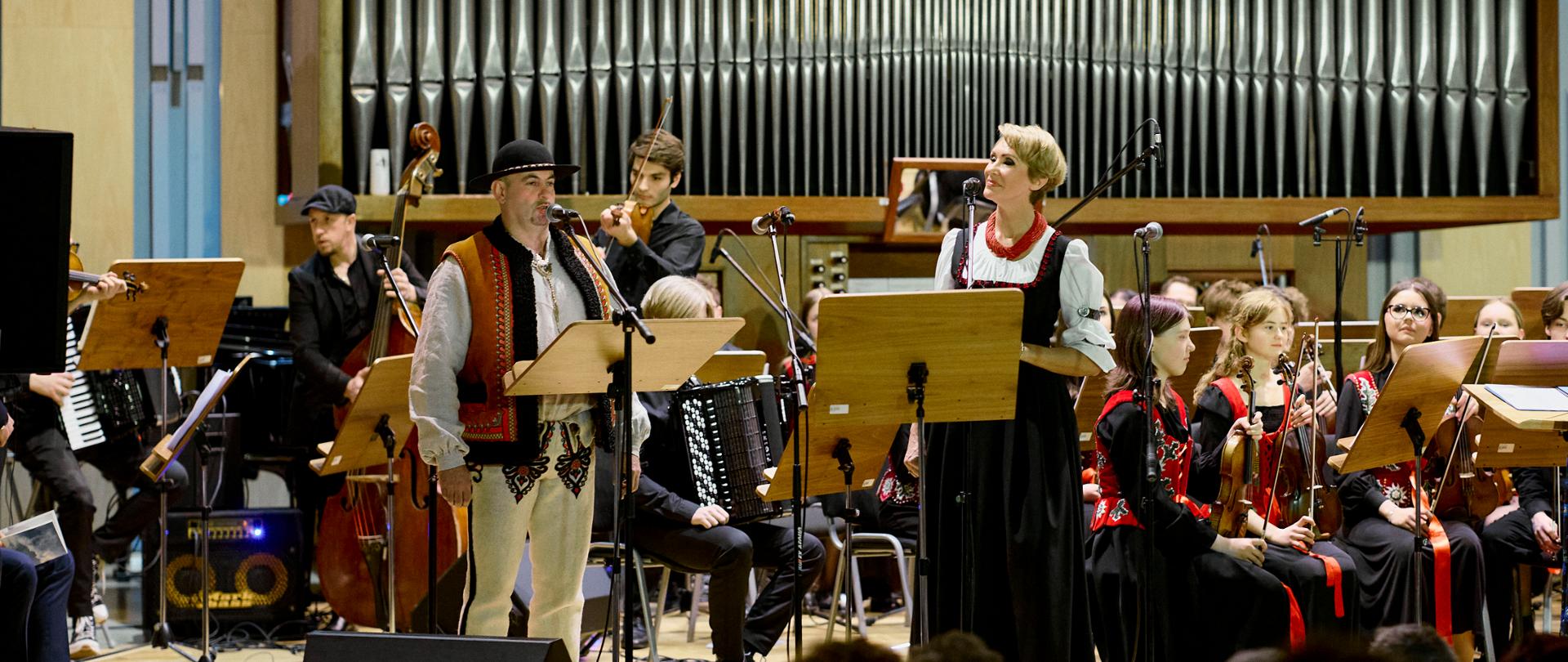 To zdjęcie z tej samej serii koncertowej, jednak tym razem na pierwszym planie obok solistki pojawia się nowy wykonawca. Oto kluczowe elementy tego ujęcia: Nowy solista: Po lewej stronie mikrofonu stoi mężczyzna ubrany w pełny strój góralski – w tym haftowane spodnie (portki), bogato zdobioną kamizelkę (cuchę) oraz charakterystyczny czarny kapelusz z muszelkami. Solistka: Obok niego występuje kobieta znana z poprzednich zdjęć, ubrana w białą bluzkę, czarną suknię i czerwone korale, co sugeruje wspólny występ w klimacie folkowym. Zespół muzyczny: W tle widoczni są instrumentaliści, w tym akordeonista, kontrabasista oraz sekcja smyczkowa. Muzycy łączą klasyczny ubiór koncertowy z elementami regionalnymi. Sceneria: Niezmiennie tło stanowią monumentalne organy, a u dołu zdjęcia widoczna jest publiczność obserwująca występ. Zdjęcie to jeszcze wyraźniej podkreśla podhalański lub góralski charakter wydarzenia, łącząc tradycję ludową z powagą sali koncertowej
