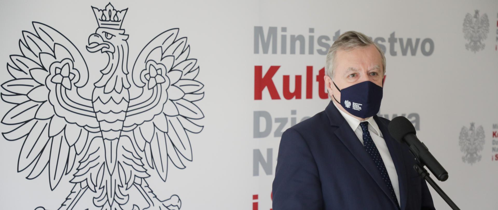 Na zdjęciu wicepremier oraz minister kultury, dziedzictwa narodowego i spotu prof. Piotr Gliński, stojący na tle ścianki w kolorze białym z godłem Polski i logo resortu. Mężczyzna ubrany w ciemny garnitur, białą koszulę i ciemny krawat w kropki. Spogląda w bok obiektywu. Stoi przed mikrofonem, ustawionym na statywie. Na twarzy ma granatową maseczkę ochronną z napisem MKDNiS