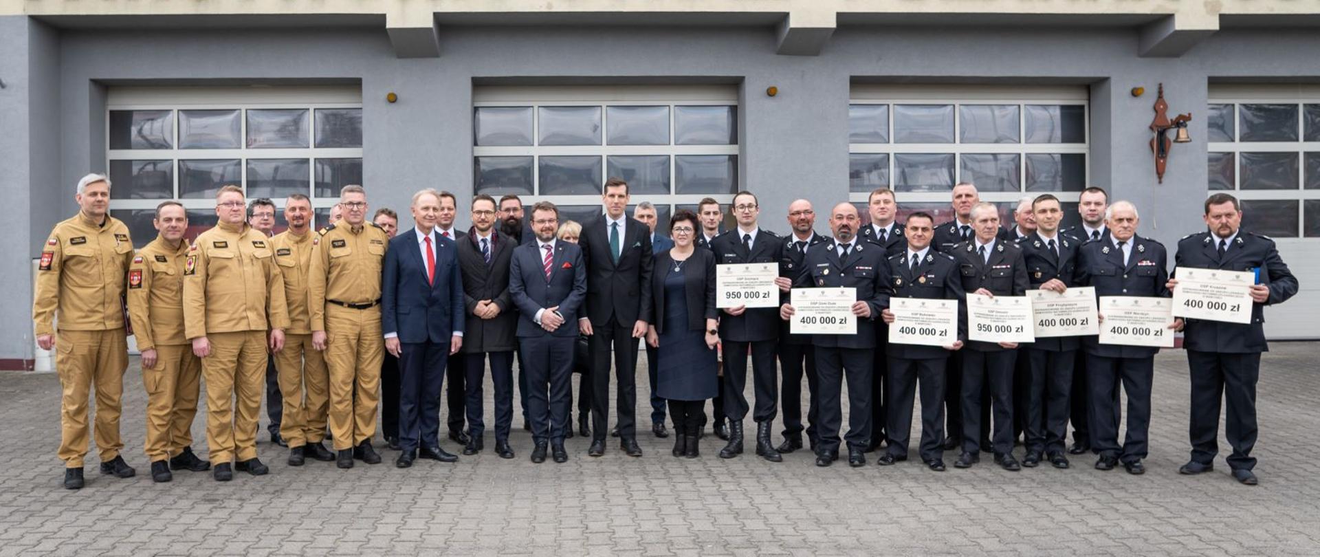 strażacy PSP, OSP, ministrowie, urzędnicy pozują do zdjęcia na tle strażnicy KP PSP powiatu łódzkiego wschodniego, strażacy OSP na pierwszym planie trzymają w rękach wręczone promesy na zakup samochodów 