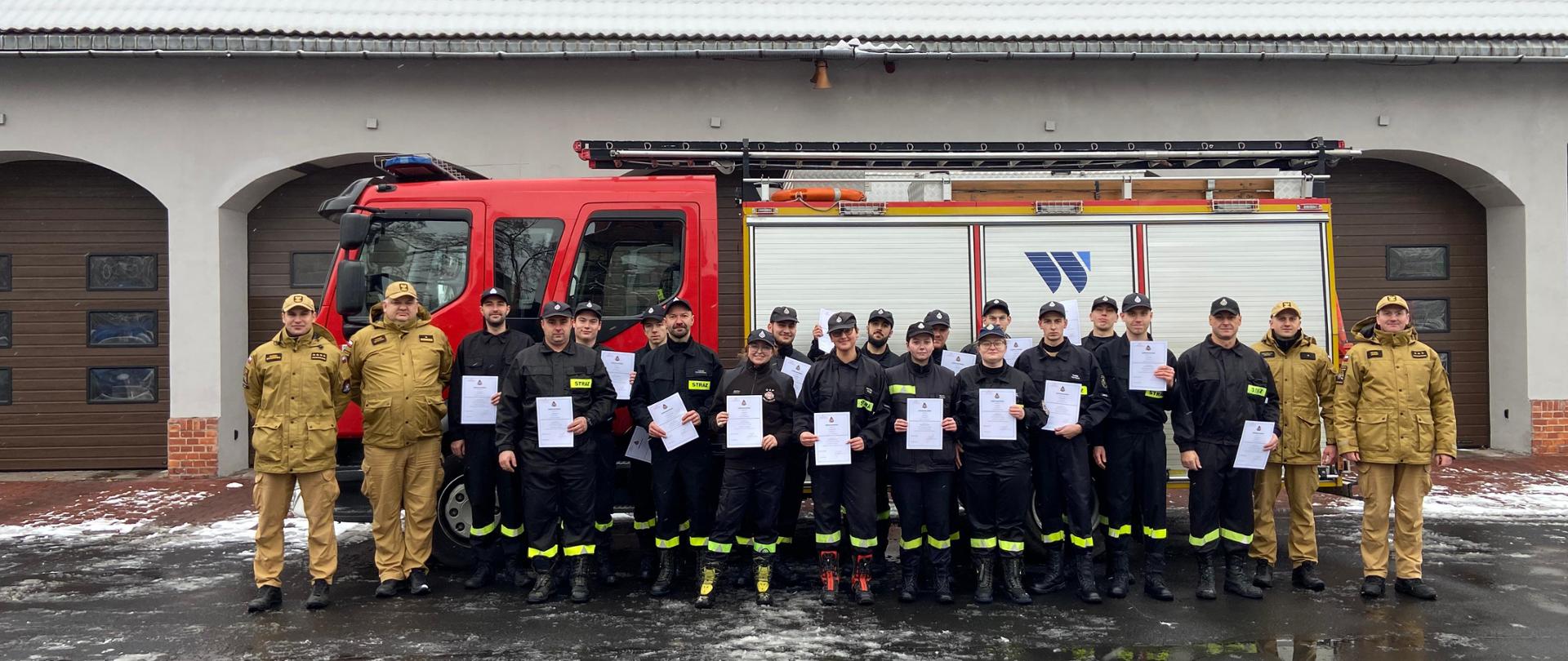 Grupa strażaków ochotników stoi przed Komendą Miejską PSP w Zabrzu wraz z kadrą egzaminacyjną oraz zastępcą komendanta PSP w Zabrzu, mł. bryg. Szymańskim. Ochotnicy trzymają w rękach zaświadczenia o nabyciu kwalifikacji strażaka ratownika OSP. W tle widoczny czerwony wóz strażacki i budynek komendy z zaśnieżonym dachem.