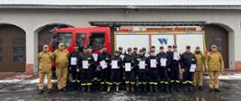 Grupa strażaków ochotników stoi przed Komendą Miejską PSP w Zabrzu wraz z kadrą egzaminacyjną oraz zastępcą komendanta PSP w Zabrzu, mł. bryg. Szymańskim. Ochotnicy trzymają w rękach zaświadczenia o nabyciu kwalifikacji strażaka ratownika OSP. W tle widoczny czerwony wóz strażacki i budynek komendy z zaśnieżonym dachem.