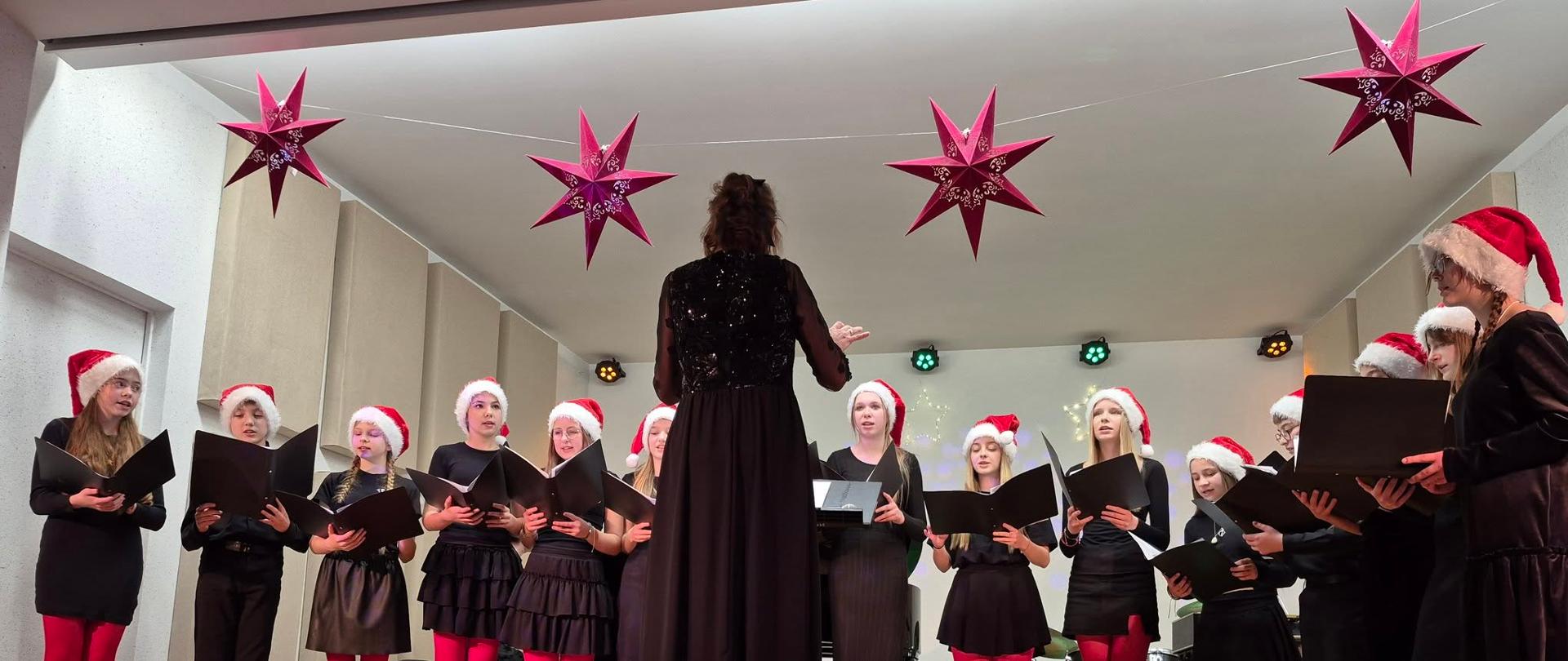 Na zdjęciu widać chór i panią dyrygentkę. Chór stoi na scenie, która jest ozdobiona świątecznymi dekoracjami. Uczniowie śpiewają, trzymają teczki, a na głowach mają mikołajkowe czapki. 