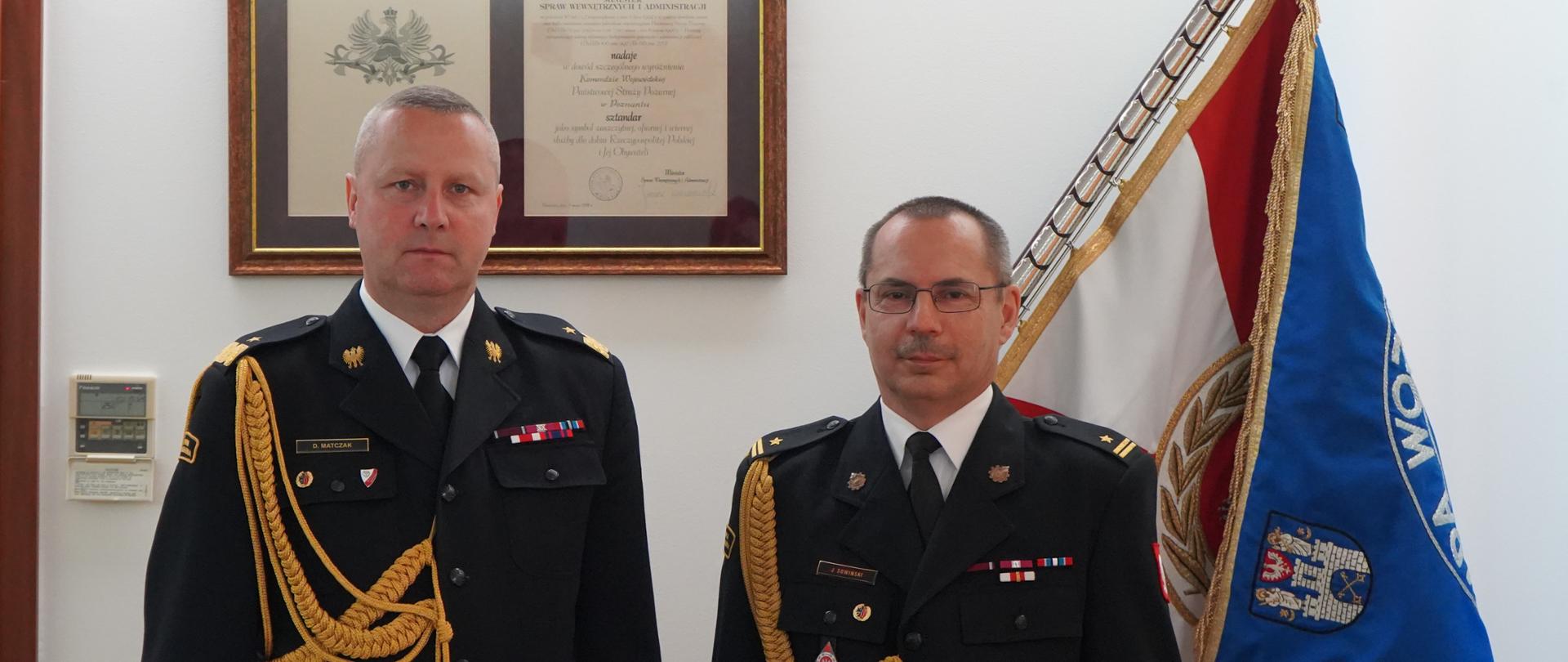 dwóch strażąków w mundurach ze sznurem stoi obok siebie, jeden trzyma w ręku teczkę, w tle sztandar