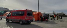 Na zdjęciu widoczny czerwony bus z sochaczewskiej Komendy Powiatowej PSP. Na dalszym planie pomarańczowy namiot, grupa uchodźców z walizkami i osoby związane z obsługą migrantów.