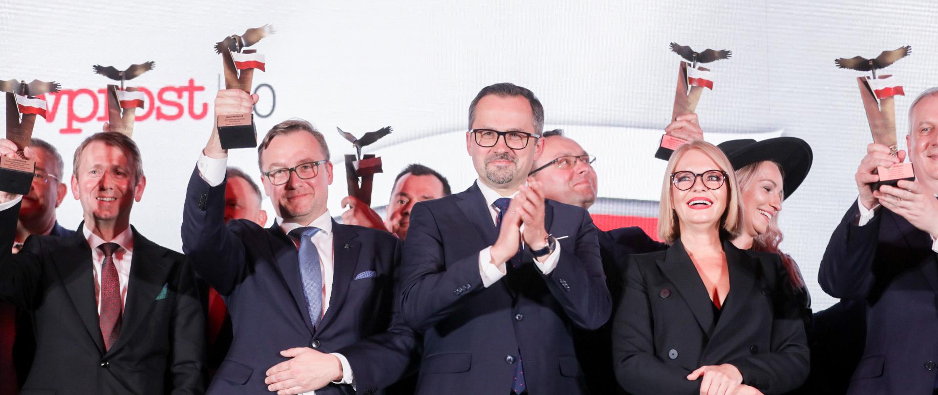 Grupa osób stoi na scenie. W środku stoi i klaszcze wiceminister Marcin Horała.