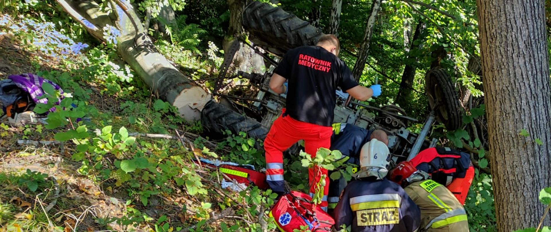 Zdjęcie przedstawia strażaków podczas udzielania kwalifikowanej pierwszej pomocy osobie poszkodowanej. Na zdjęciu znajduje się przewrócony ciągnik oraz pracujący strażacy a także ratownicy medyczni. 