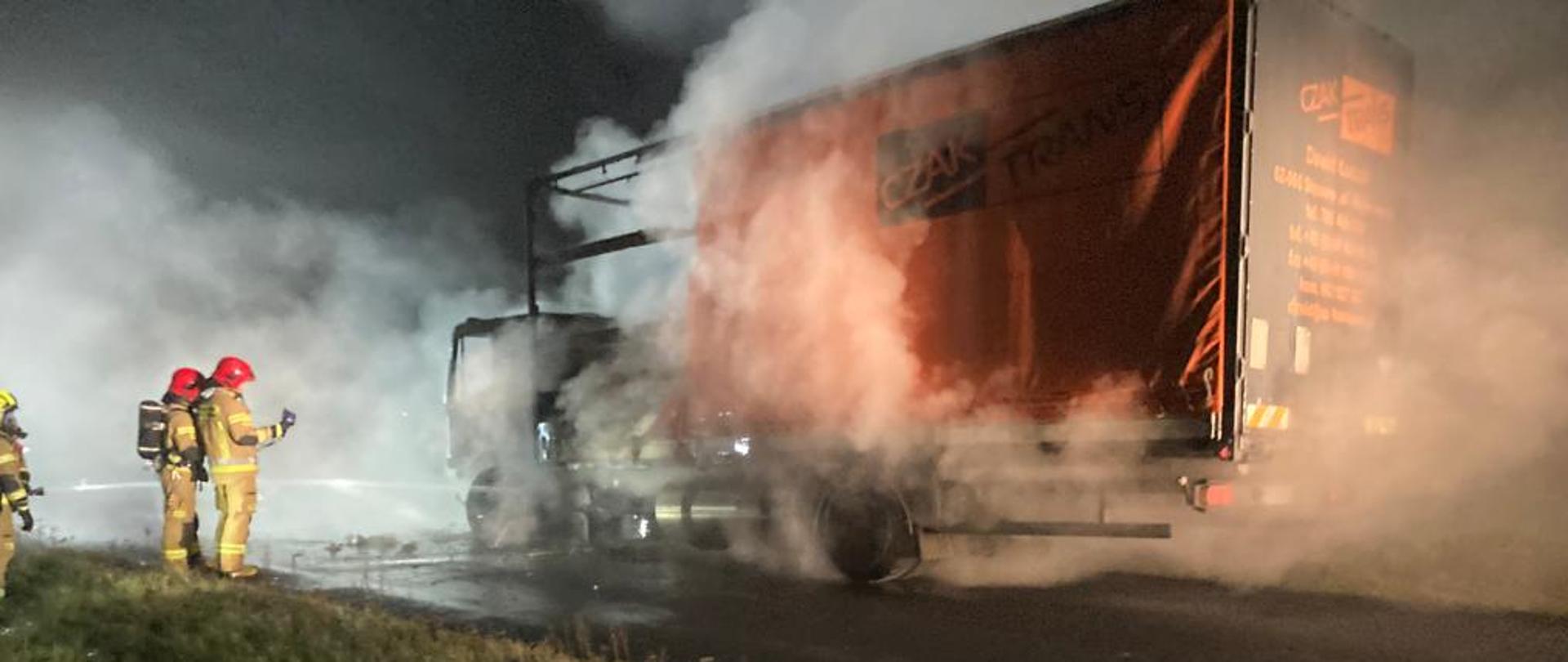 Na zdjęciu ratownicy ubrani w ODO i ubranie specjalne mierzą temperaturę spalonego pojazdu przy pomocy kamery termowizyjnej