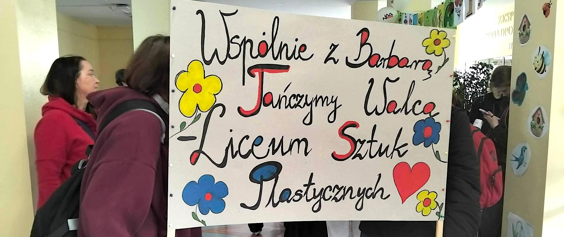 W holu szkoły stoi grupa uczniów, na pierwszym planie dwie uczennice trzymające Baner z napisem wspólnie z Barbarą tańczymy walca – liceum Sztuk Plastycznych