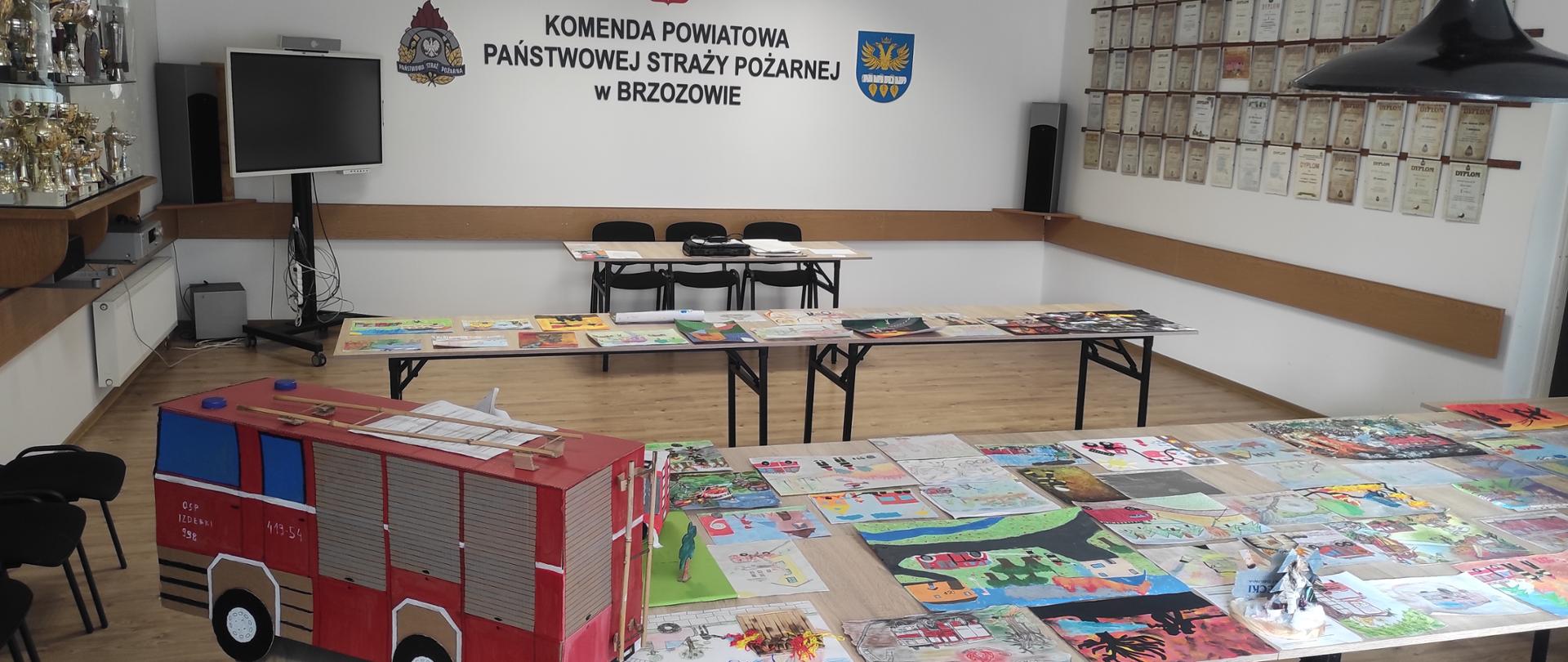Kolorowa fotografia wykonana w pomieszczeniu w pogodny dzień. Przedstawia rozłożone prace do oceny w konkursie plastycznym w pomieszczeniu świetlicy KP PSP w Brzozowie. W tle ściana z napisem Komenda Powiatowa PSP w Brzozowie.