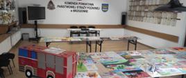  Kolorowa fotografia wykonana w pomieszczeniu w pogodny dzień. Przedstawia rozłożone prace do oceny w konkursie plastycznym w pomieszczeniu świetlicy KP PSP w Brzozowie. W tle ściana z napisem Komenda Powiatowa PSP w Brzozowie.