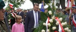 Minister Czarnek niesie duży wieniec biało-czerwonych kwiatów, obok niego mała dziewczynka w różowej bluzie.