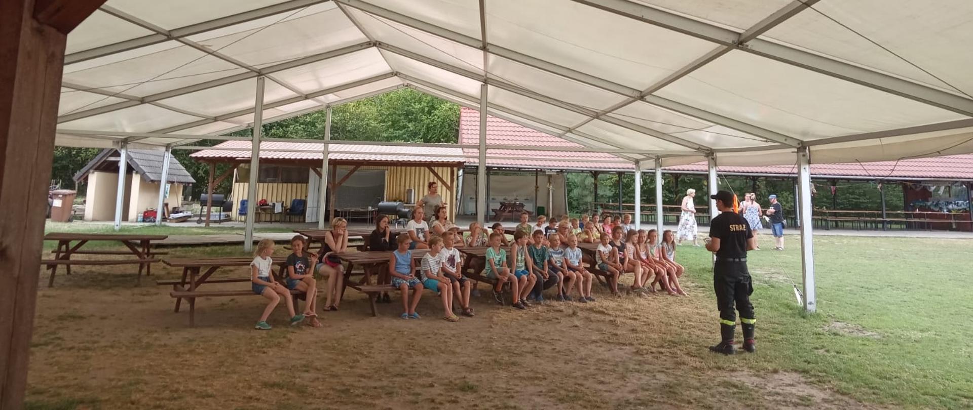 Na zdjęciu pod namiotem siedzą dzieci na ławkach a przed nimi strażak prowadzi pogadankę na temat bezpieczeństwa 