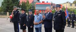 Osoby przewidziane do wyróżnienia stoją obok siebie za nimi stoi samochód pożarniczy oraz poczet sztandarowy ochotniczej straży pożarnej.