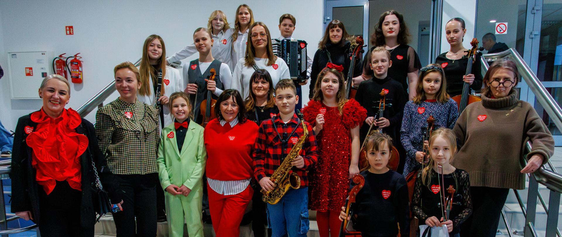 Grupa kilkunastu osób, dorosłych i dzieci stoją na schodach auli CP, niektórzy trzymają w rękach instrumenty muzyczne, wszyscy mają przyklejone do ubrań serduszka WOŚP.