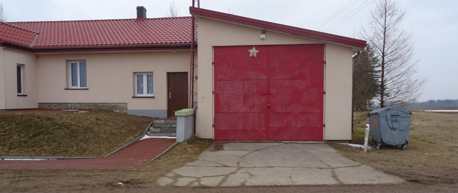 Budynek murowany jasno żółty. Brama garażowa koloru czerwonego. dach koloru czerwonego. Podjazd do bramy garażowej wylany asfaltem. 