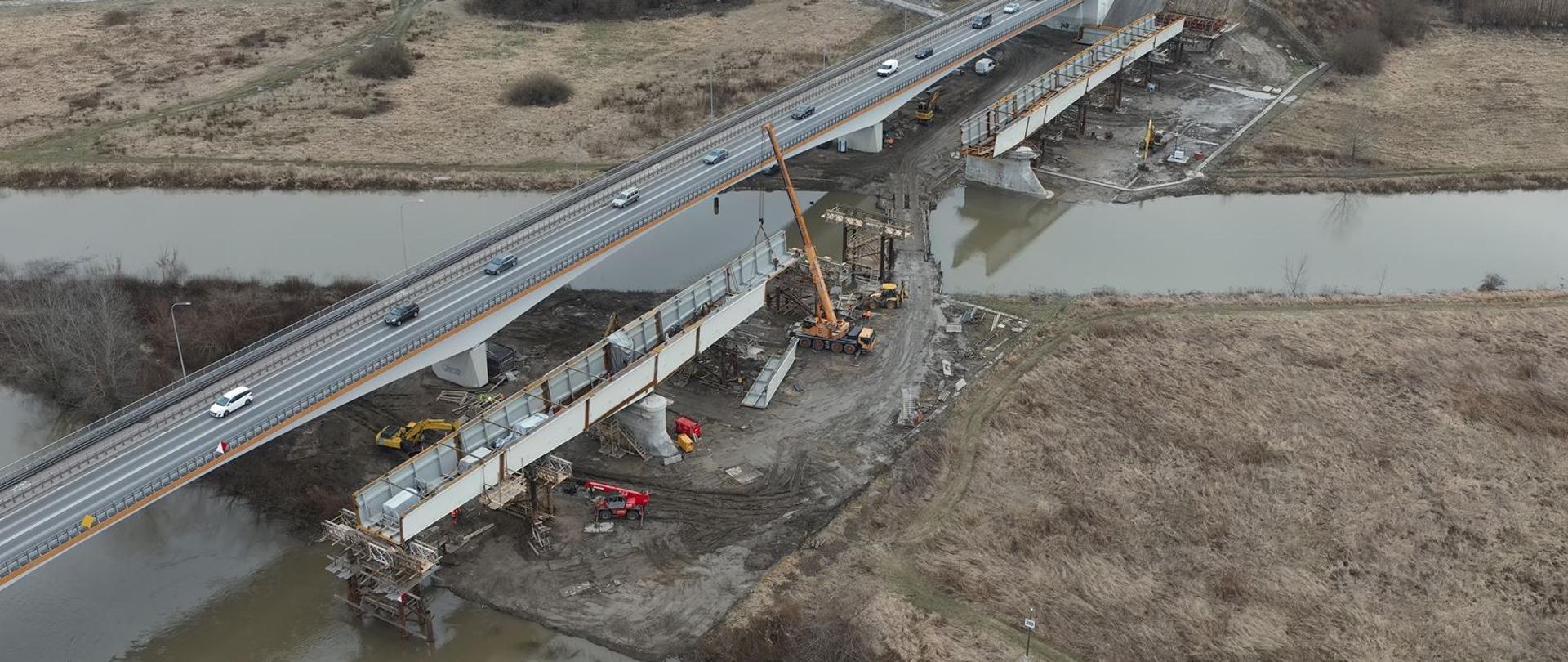 Budowa mostu w Sandomierzu - ujęcie z góry - rzeka, teren przybrzeżny, istniejący most, po którym jadą samochody, obok budowa konstrukcji nowego obiektu, na podporach zamontowane elementy stalowe, dźwigi i koparki 