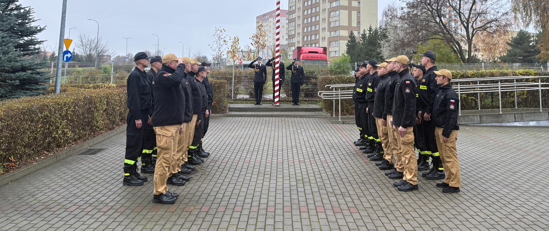 Strażacy stoją na placu przed jednostką w dwóch szeregach na przeciw siebie. Przy maszcie stoją strażacy i oddają hołd. Na maszcie wisi flaga państwowa. 