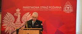 Funkcjonariusz PSP stoi za drewnianą mównicą z emblematem Państwowej Straży Pożarnej. Pochyla się lekko nad mikrofonem, czytając przygotowane wystąpienie. W tle znajduje się czerwona ścianka z godłem państwowym oraz napisem „Państwowa Straż Pożarna”, co podkreśla oficjalny charakter wydarzenia.
