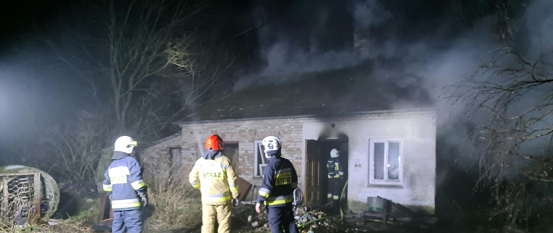 Zdjęcie przedstawia trzech strażaków znajdujących się przed palącym budynkiem. Z budynku wydobywają się kłęby dymu. 