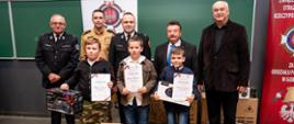 Zajęcie przedstawia laureatów trzech pierwszych miejsc w eliminacjach powiatowych Ogólnopolskiego Turnieju Wiedzy Pożarniczej w powiecie gorlickim z nagrodami i dyplomami oraz gości wręczających nagrody rzeczowe i dyplomy.