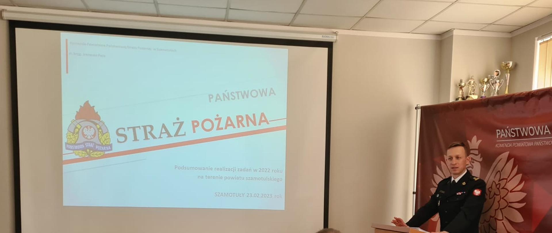 strażak stoi przy mównicy, przemawia, obok duży ekran ze slajdem na którym jest logo psp oraz napis straż pożarna