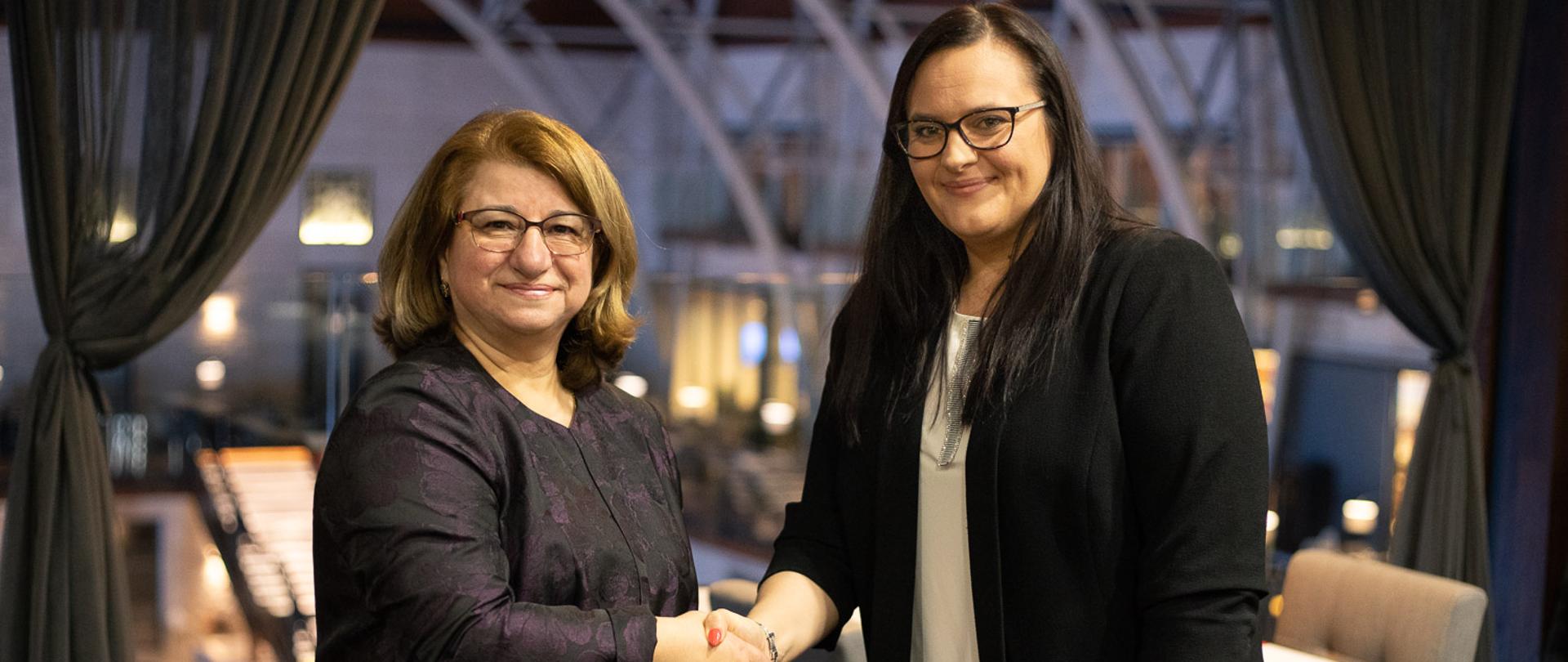 Dwie kobiety podają sobie ręce. Stoją na tle stolików z krzesłami.