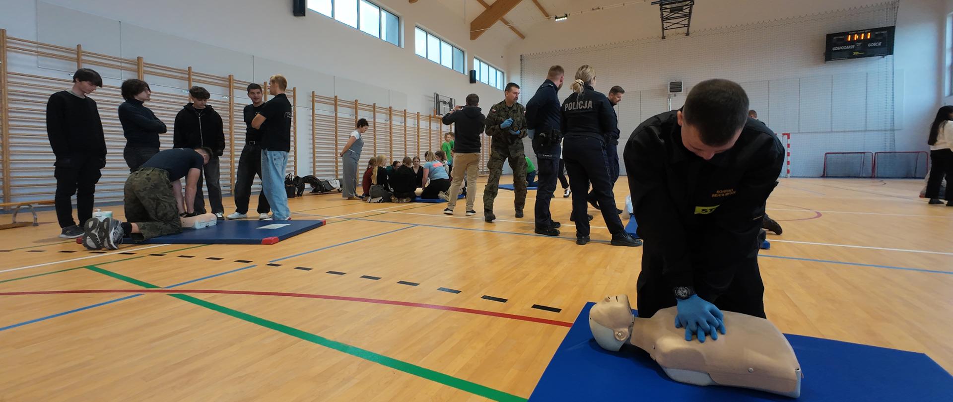 Na zdjęciu widoczny strażak oraz uczniowie wykonujący masaż serca na manekinach ćwiczebnych.