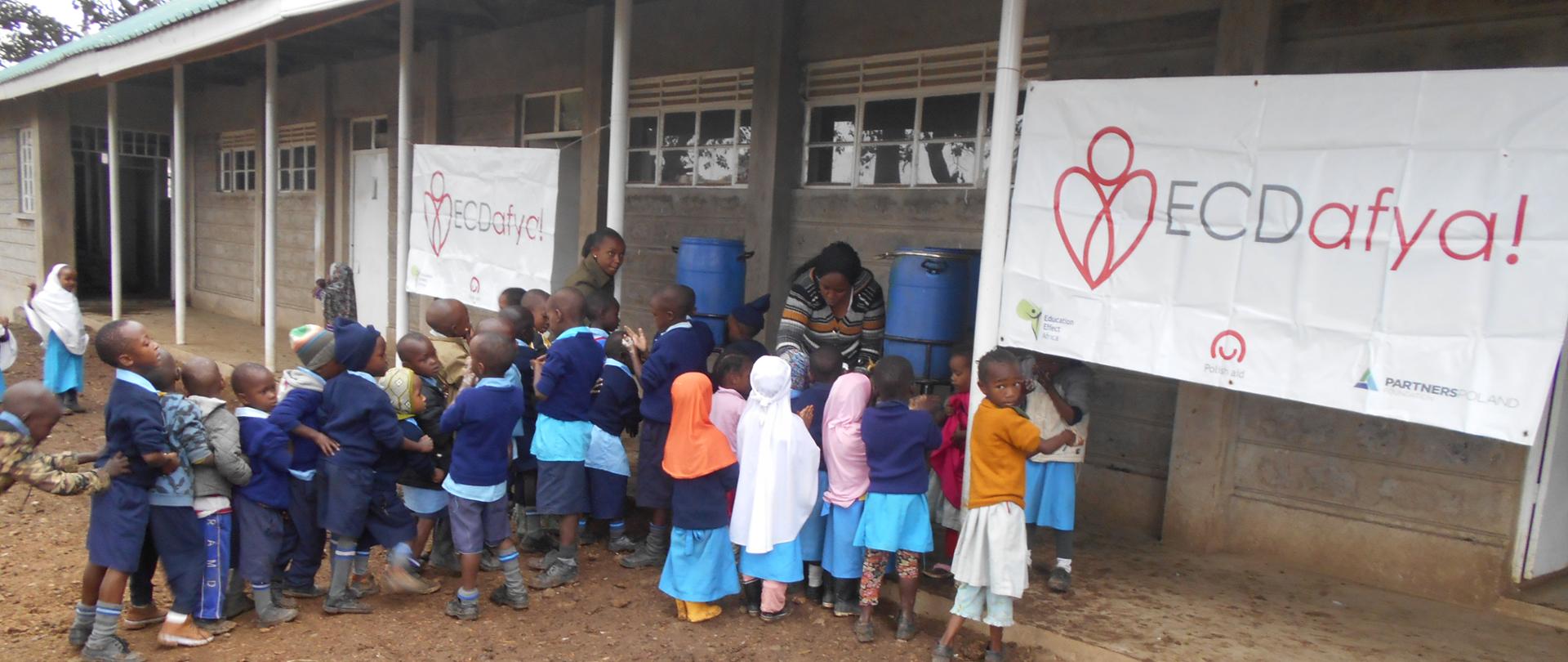 Grupa dzieci przed budynkiem myje ręce w wodzie z beczek