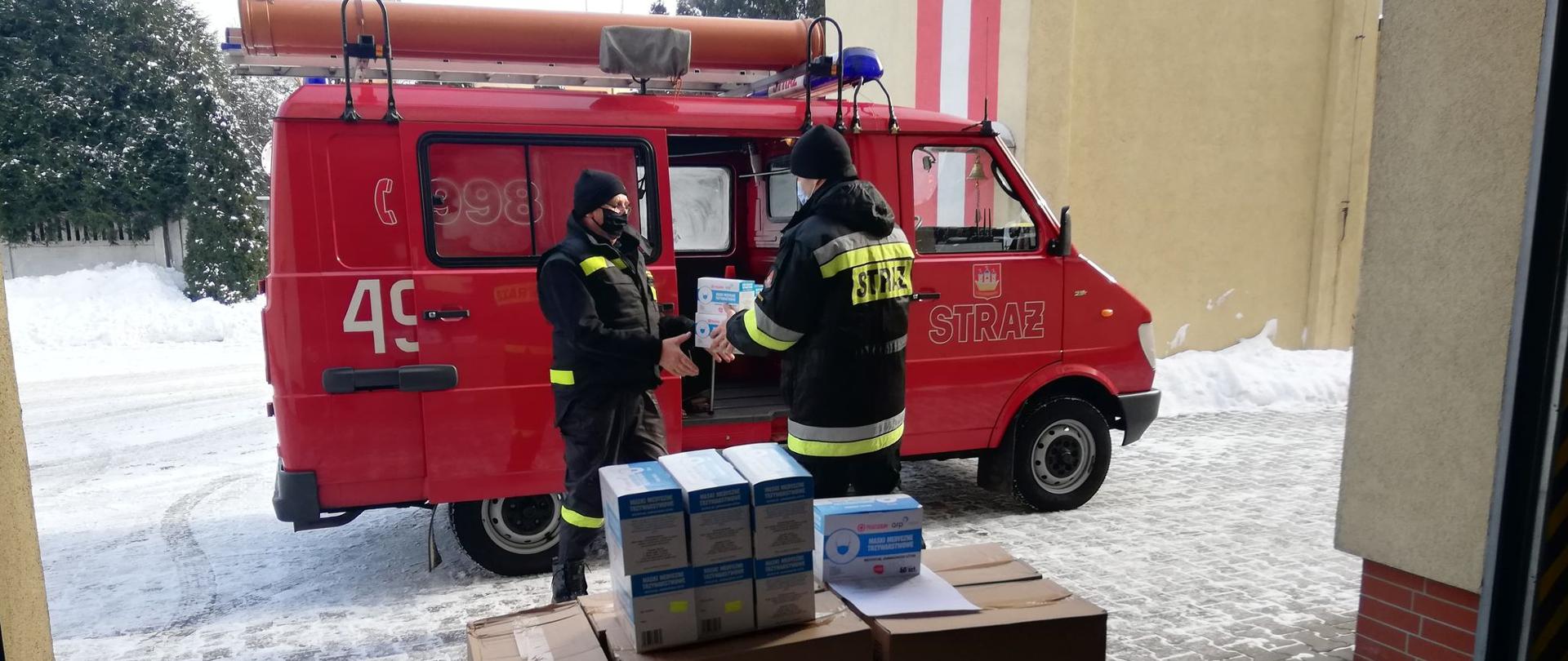 Funkcjonariusze przekazują środki ochrony indywidualnej przedstawicielom jednostek OSP wspierającym narodowy program szczepień. Funkcjonariusz wręcza karton ze środkami, który odbiera druh osp i przenosi do samochodu. Pojazd zapakowany na placu KP PSP. Całość środków przekazywanych na daną gminę znajduje się w garażu.