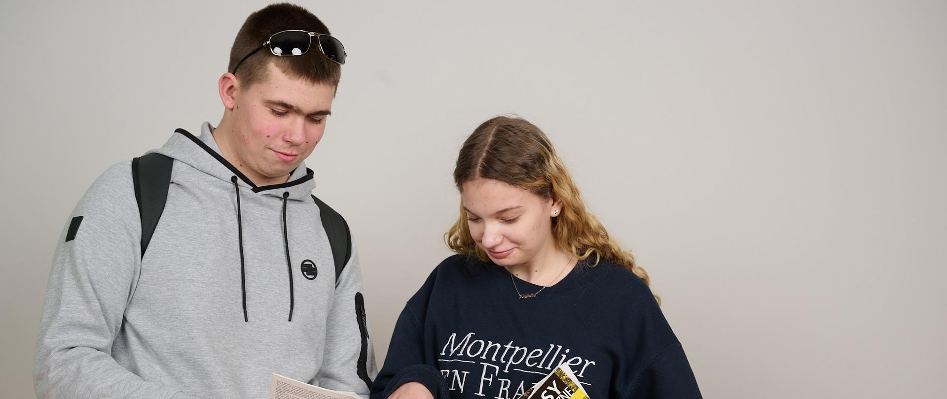 Dziewczyna i chłopak spoglądają na mapę, którą chłopak trzyma w ręku. Dziewczyna prawą ręką wskazuje na mapę a w lewej trzyma kolejną mapę turystyczną.